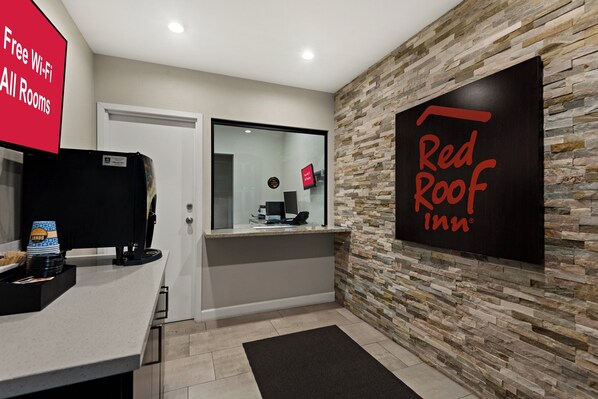 Un área de recepción con un letrero de Red Roof Inn, una puerta de vidrio, una mesa de recepción con una máquina de café y un cartel que ofrece Wi-Fi gratuito en todas las habitaciones.