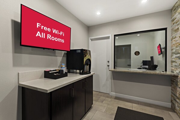 Un moderno vestíbulo de hotel con un televisor de pantalla plana que muestra “WiFi gratis en todas las habitaciones”, una cafetera y un mostrador con un espejo que refleja la sala.