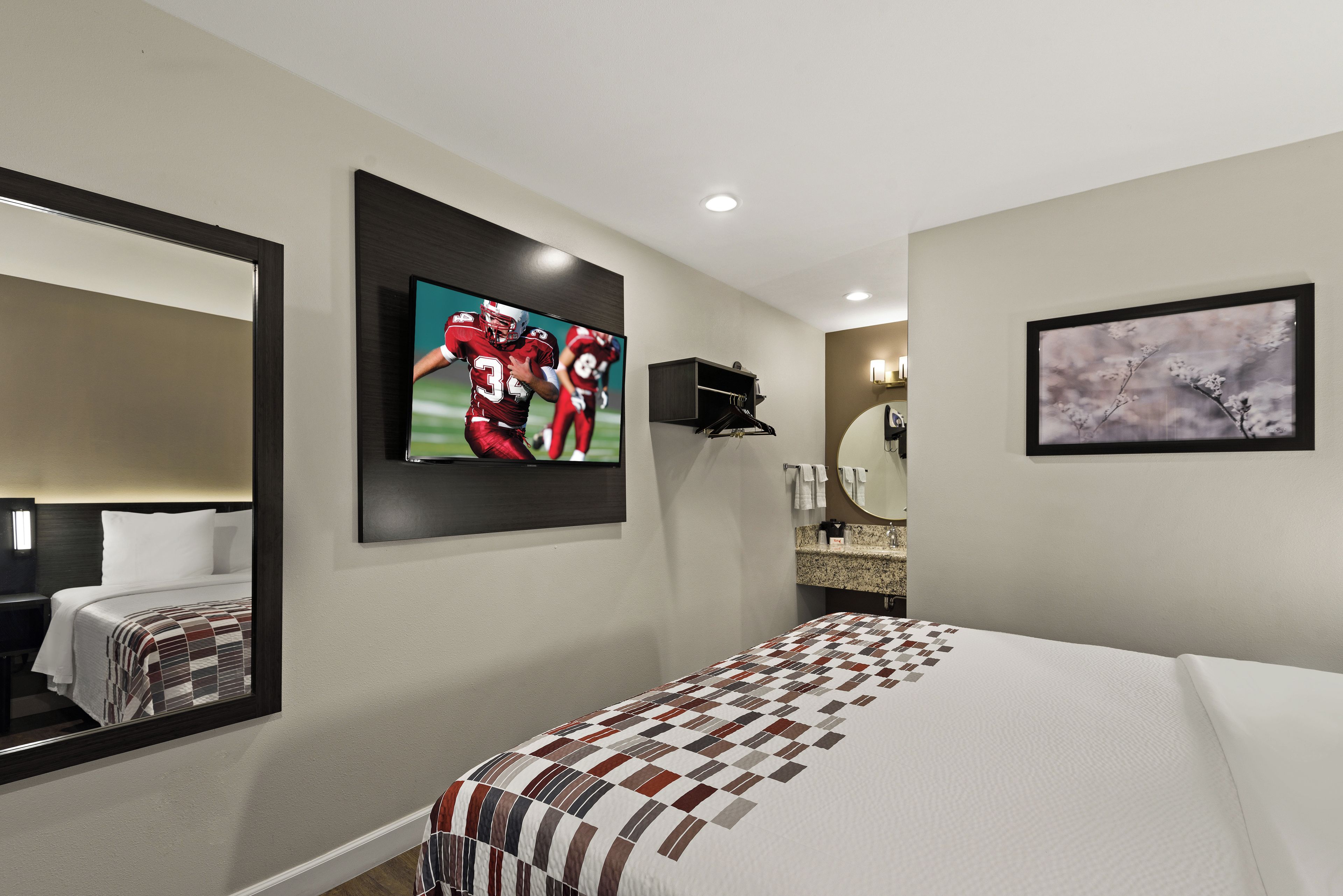 Ein Hotelzimmer mit einem großen Bett, einem an der Wand montierten Fernseher, einem Spiegel, einem Gemälde und einer kleinen Ablage mit Pflegeprodukten.