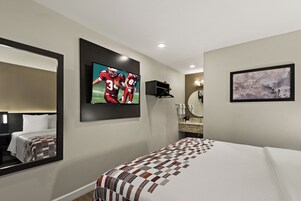 A hotel room with a large bed, a TV mounted on the wall, a mirror, a painting, and a small shelf with toiletries.