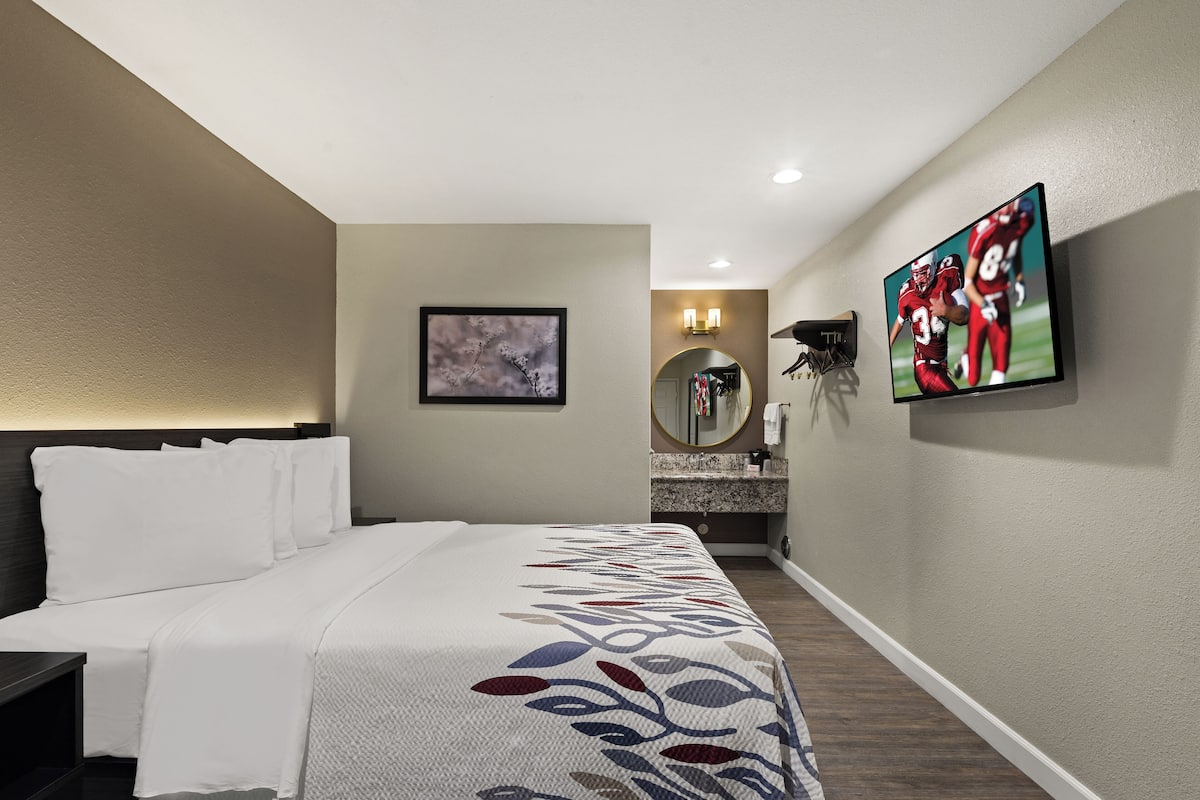 A hotel room with a bed, a TV mounted on the wall, a mirror, and a small bathroom area with a sink and toilet.