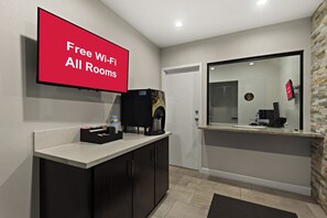 Un moderno vestíbulo de hotel con un televisor de pantalla plana que muestra “WiFi gratis en todas las habitaciones”, una cafetera y un mostrador con un espejo que refleja la sala.