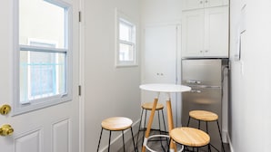 A compact kitchen with white cabinets, a stainless steel refrigerator, a round white table, and two wooden stools.
