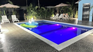 An outdoor swimming pool with lounge chairs and umbrellas, surrounded by tropical plants.