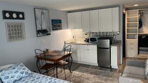 Cocina compacta con armarios blancos, microondas y refrigerador. Hay un comedor con mesa y sillas, y una cama con cubrecama de estampado azul.