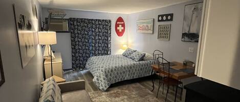 A compact room with a bed, a desk, a chair, and a sofa. There is a red emergency exit sign on the wall.