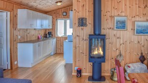 A cozy cabin interior with a central wood-burning stove, a kitchen with white cabinets, and a dining area with a table and chairs. - Gorgeous home in Noresund with WiFi (Noresund)