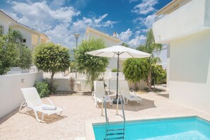 A poolside area with white lounge chairs and an umbrella, surrounded by greenery and a clear blue sky. - Protaras Delfini Villa Dv8 (Paralimni)