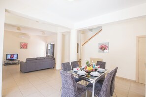 A dining area with a table set for four, surrounded by patterned chairs. A sofa and a television are visible in the background.