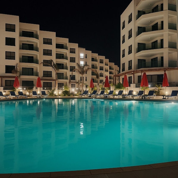 A nighttime view of a hotel pool area with multiple levels of balconies, red umbrellas, and lounge chairs.