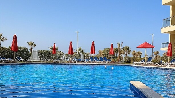A swimming pool with clear blue water, surrounded by lounge chairs and red umbrellas. - Find Your Holiday Haven at Porto Said Resort Chalets (Al-Manasrah)