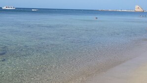 A beach with clear shallow waters, a few boats, and a distant rocky outcrop.