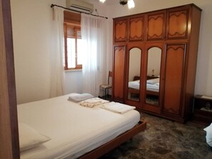 Un dormitorio con cama, armario, mesita y ventana con cortinas.