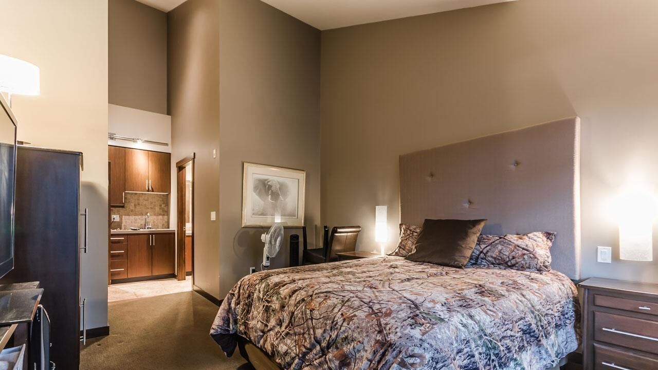 A bedroom with a large bed, a desk, a chair, a dresser, and a kitchenette in the background.