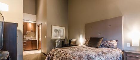 A bedroom with a large bed, a desk, a chair, a dresser, and a kitchenette in the background.