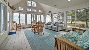 A spacious room with a large window overlooking a lake, a wooden dining table with chairs, a grey sofa, and a patterned rug. - Spacious, Peaceful Lake House w/ Sandy Beach (Northport)