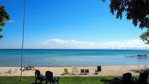 A beach with clear blue water, a wooden pier, and several beach chairs and tables. - Spacious, Peaceful Lake House w/ Sandy Beach (Northport)