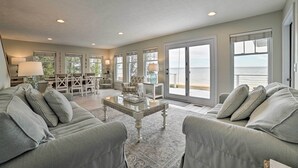 A spacious living room with a large glass coffee table, grey upholstered sofas, and a view of the ocean through large sliding doors. - Spacious, Peaceful Lake House w/ Sandy Beach (Northport)