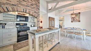 A kitchen with a brick wall, modern appliances, and a dining area with white chairs and a wooden table. - Waterfront Suttons Bay: Wine Trail, Pickleball Ct (Suttons Bay)