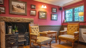 A room with a fireplace, two upholstered chairs, a small table, and framed photographs on the wall.