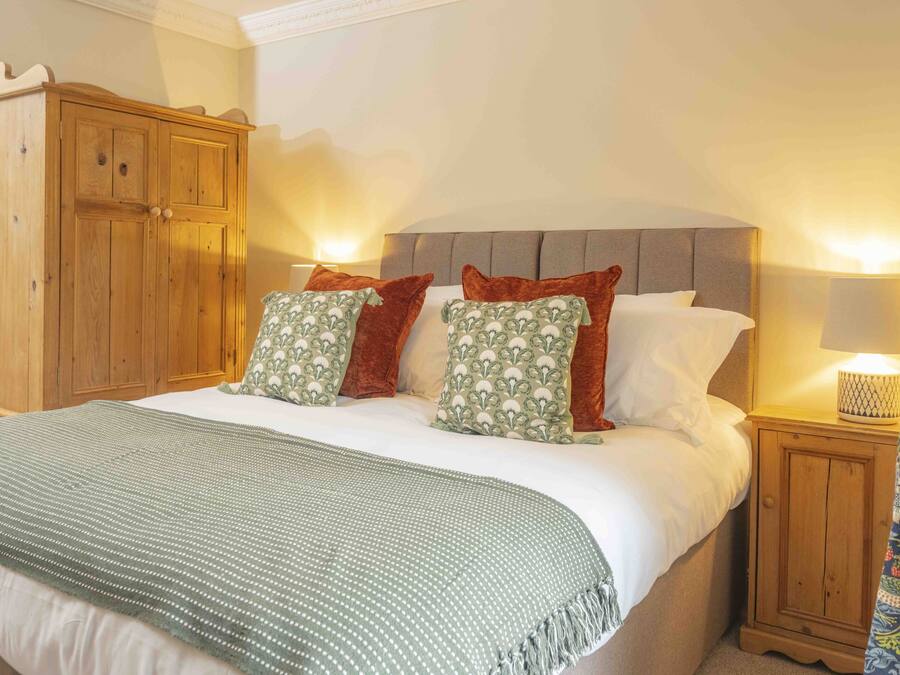 A neatly made bed with a green blanket, two pillows with a floral pattern, and a grey headboard. There is a wooden wardrobe to the left and a nightstand with a lamp to the right.