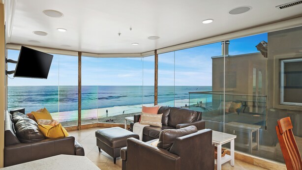 A modern living room with a large glass wall overlooking the ocean, furnished with leather sofas and a flat-screen TV. - Oceanfront Penthouse: Luxury Views, A/C & Parking! (San Diego)