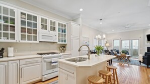 A modern kitchen with white cabinetry, a central island with stools, and a dining area with a view. - 110 Waters Edge! Luxury River Views with Community Pool, Screen Porch (Folly Beach)