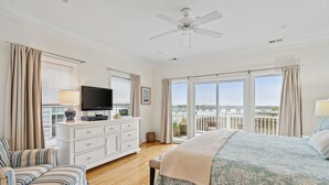 A bedroom with a large bed, a television on a dresser, a chair, and a balcony with a view. - 110 Waters Edge! Luxury River Views with Community Pool, Screen Porch (Folly Beach)