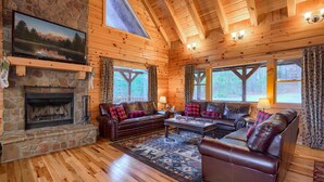 A cozy cabin interior with a stone fireplace, wooden walls, leather sofas, and a large window overlooking a forest. - Hidden Lake Lodge: Lake + 30 Acres Hocking Hills (Logan)