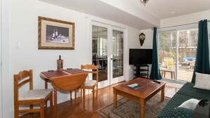 Un salon comprenant une table à manger en bois, deux chaises, un canapé, une table basse, un téléviseur et une grande fenêtre avec des rideaux.