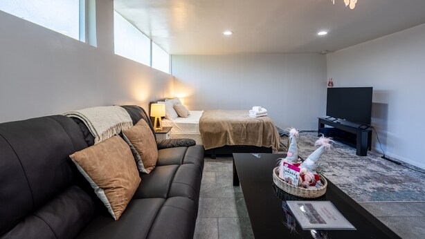 Une chambre d’hôtel moderne avec un canapé en cuir noir, un lit recouvert d’une couverture beige, un téléviseur et un lustre.
