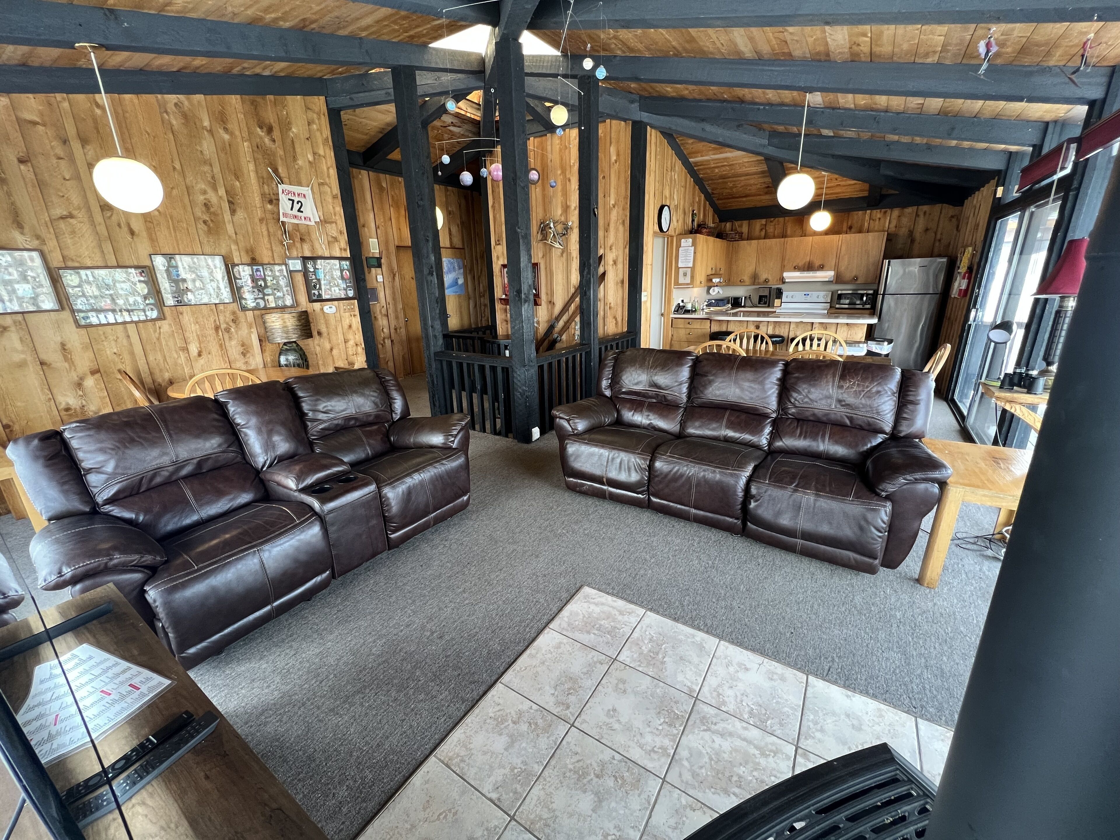 A cozy living room with two leather sofas, a wooden ceiling, and a kitchen area in the background.
