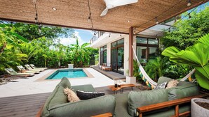 Un área junto a la piscina con cubierta de madera, asientos acolchados verdes, una hamaca y exuberante vegetación tropical.