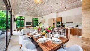 Un comedor moderno con una mesa de madera dispuesta para seis personas, rodeada de sillas blancas, en una habitación espaciosa con techo de madera y grandes ventanales con vistas a la vegetación.