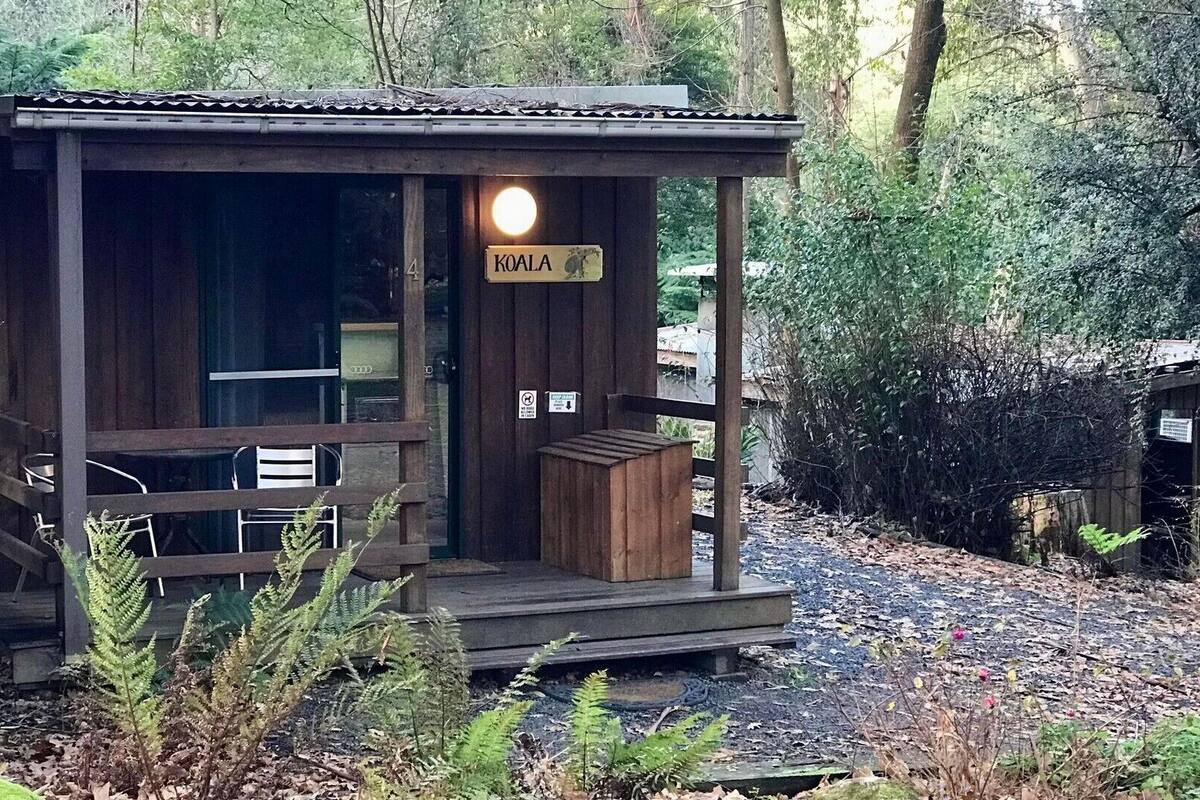 A small wooden cabin with a porch, a sign reading "KOALA", and a trash bin.