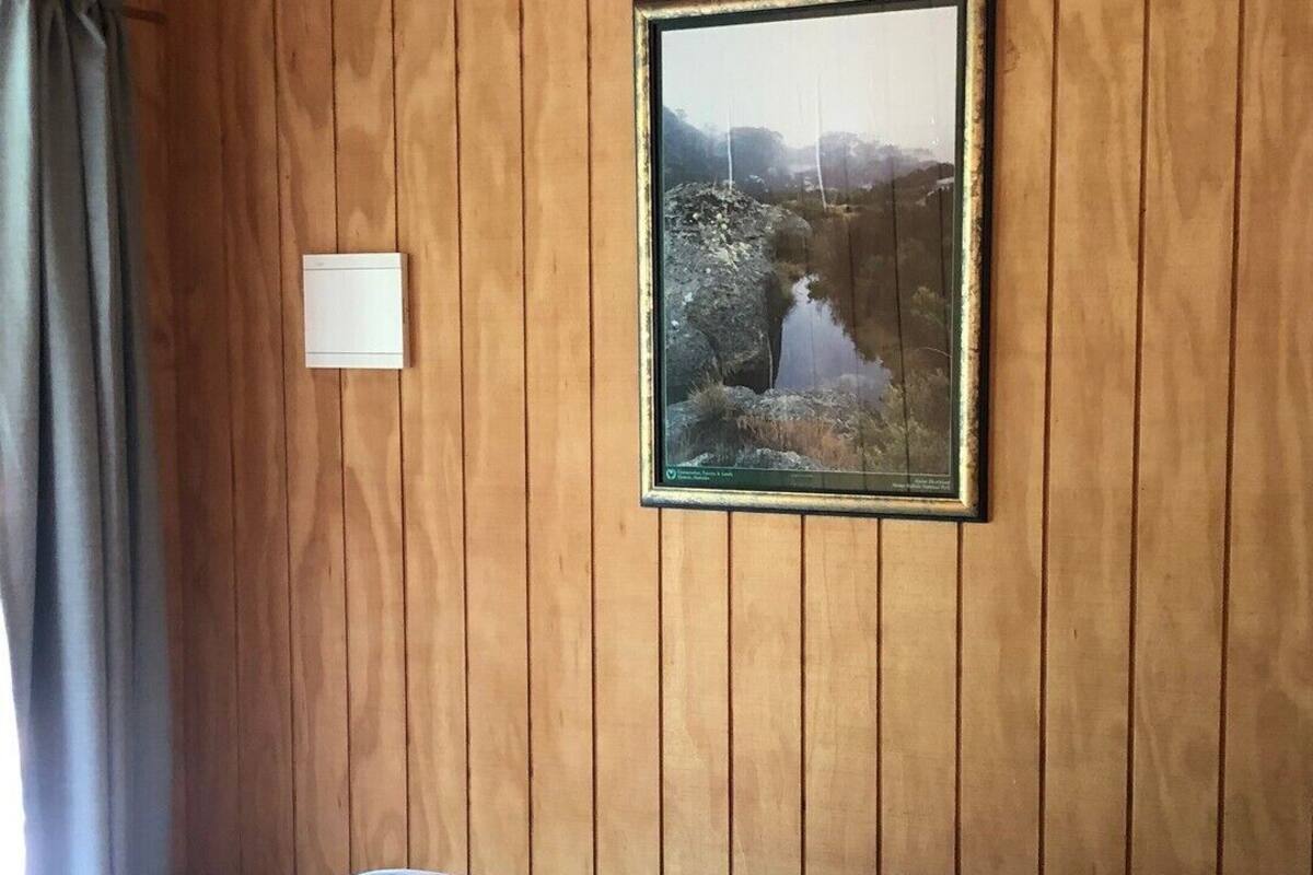 A room with wooden panel walls, a bed with a floral quilt, a framed picture of a landscape, and a light switch on the wall.
