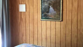 Una habitación con paneles de madera, una cama con colcha floral, un cuadro enmarcadosi un paisaje y un interruptor de luz en la pared.