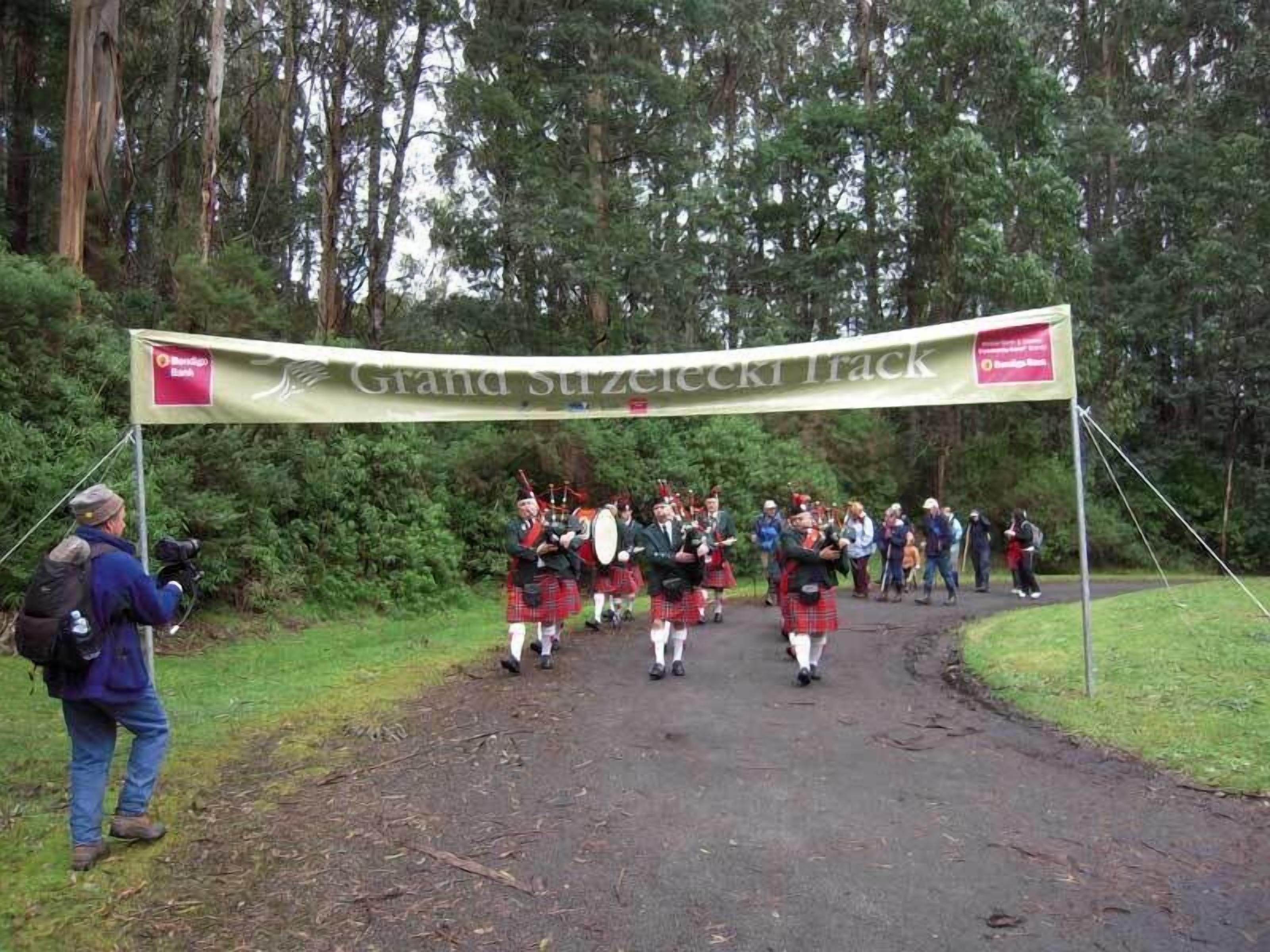 A group of people in kilts and bagpipes walking under a banner that reads "Grand Szuzelecki Track".