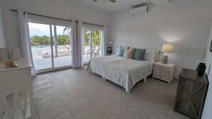 A bedroom with a bed, a nightstand, a ceiling fan, and a view of the beach through large windows.