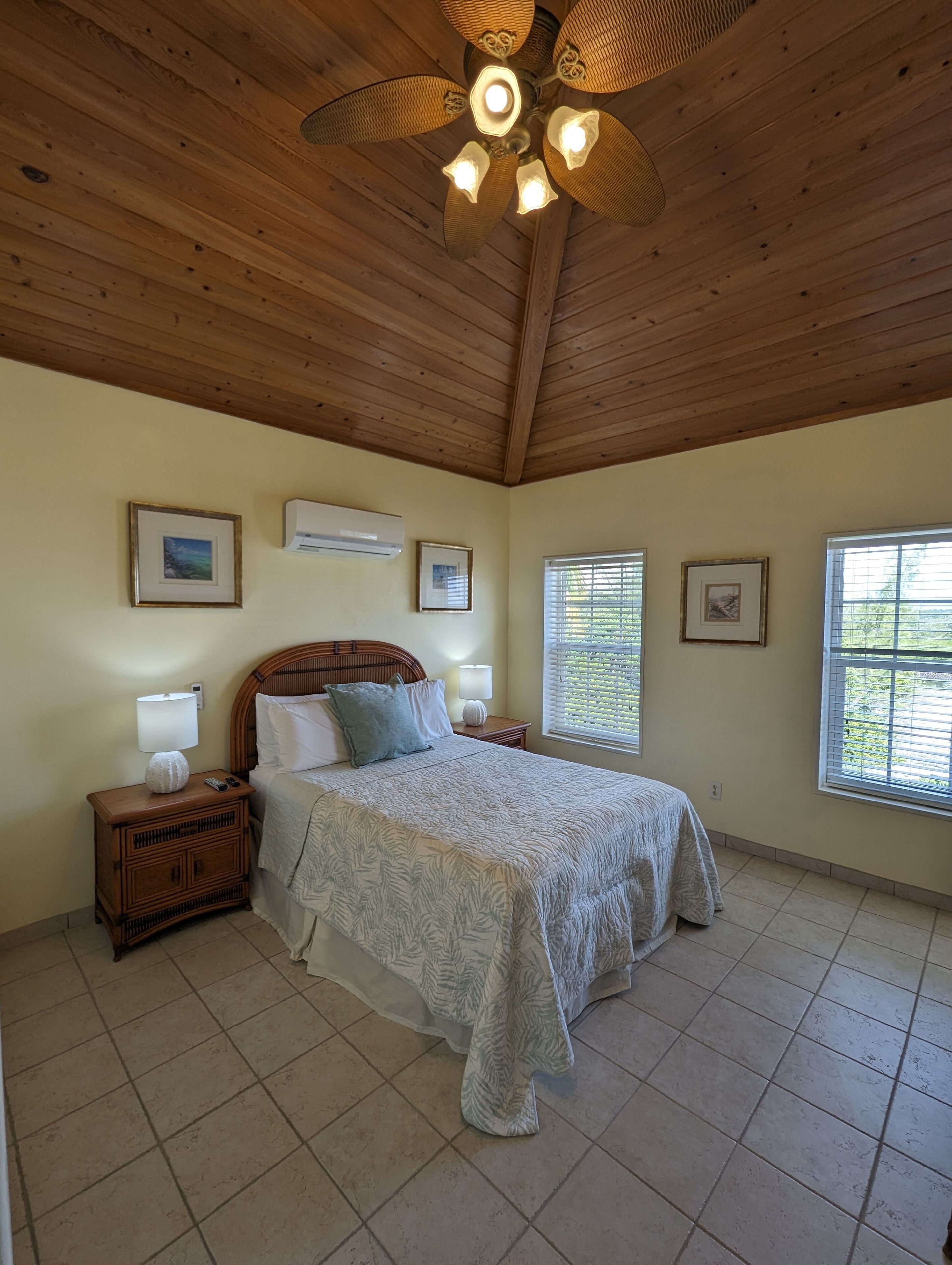 A bedroom with a wooden ceiling, a bed with a patterned bedspread, a nightstand with a lamp, and framed pictures on the wall.
