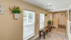 A hallway with beige walls, wooden flooring, and a bench. There are two windows with blinds, a potted plant, and a wall-mounted light fixture. - StockYards! Less than 4 minutes-Sleeps 8-Lee House, TCU close by (Fort Worth)