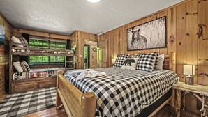 Quarto estilo cabana com beliche, uma cama grande com roupa de cama xadrez, cabeceira de madeira, mesa de cabeceira e uma imagem de urso pendurada na parede.