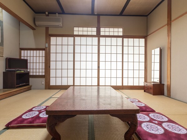 A traditional Japanese room with tatami flooring, a tatami mat table, sliding shoji doors, a television on a low shelf, and a wooden cabinet.