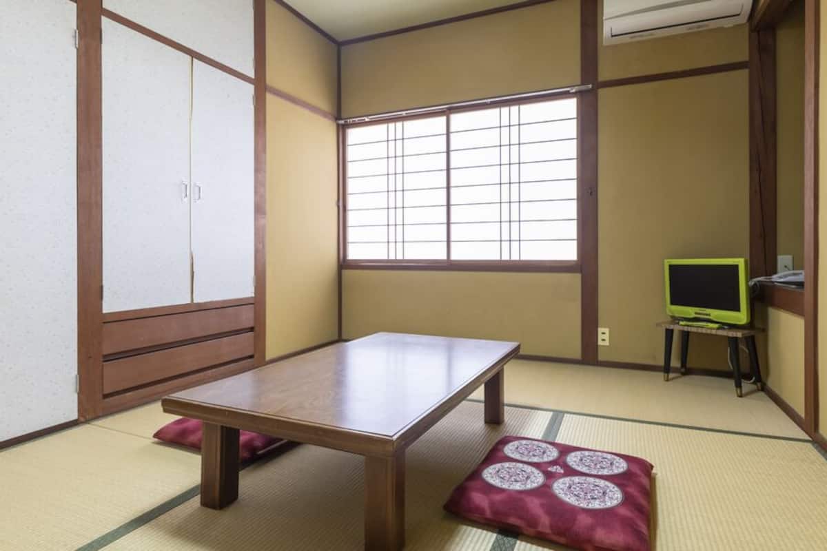 Une pièce de style japonais traditionnel, avec un sol recouvert de tatamis, une table en bois, un téléviseur posé sur un petit meuble et des portes coulissantes.