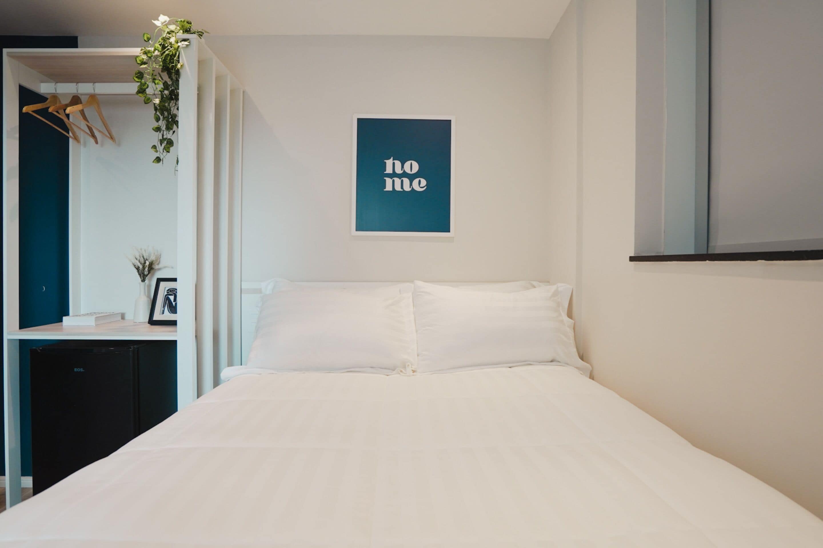 A bedroom with a bed, a small desk with a plant, a mirror, and a framed picture.