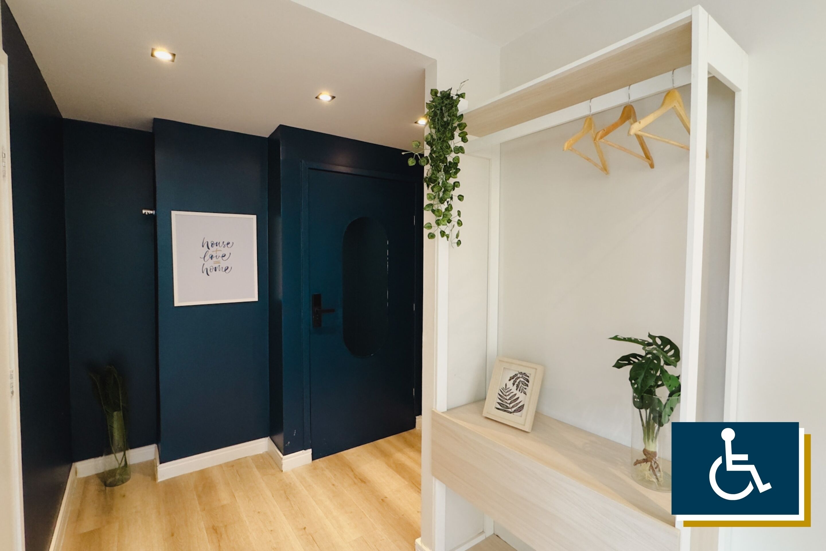 Um corredor com paredes azul-escuro, soalho de madeira, uma porta com uma placa que diz “house love home” e uma estante branca com uma planta e um quadro emoldurado.