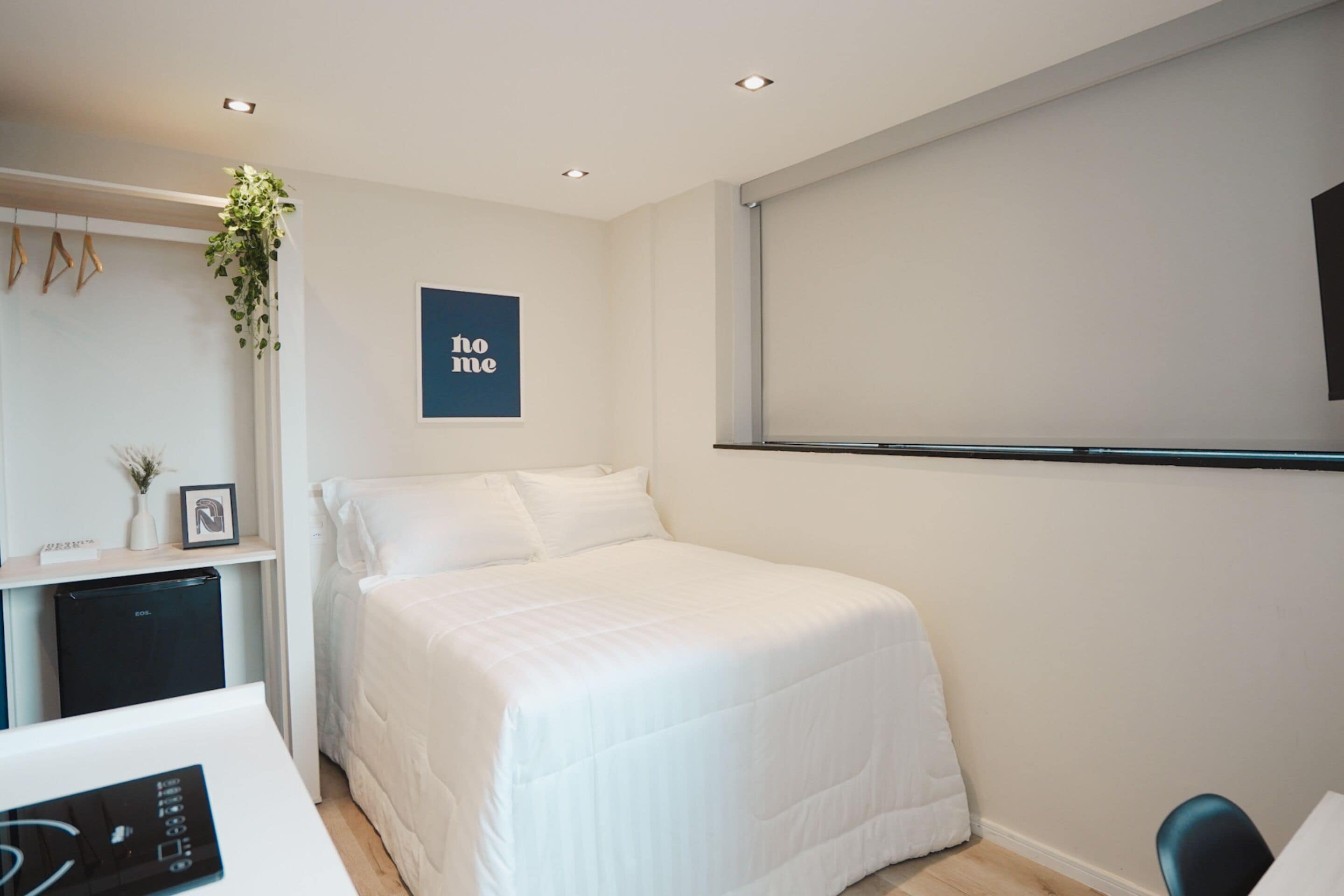 A modern hotel room with a neatly made bed, a desk with a computer, a small fridge, and a wall-mounted TV.
