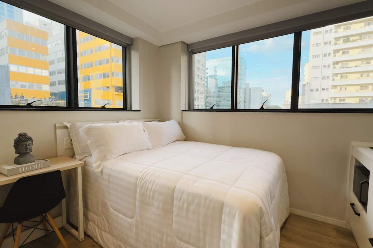 Un dormitorio con una cama, un escritorio y una silla. Amplias ventanas con vista a la ciudad.