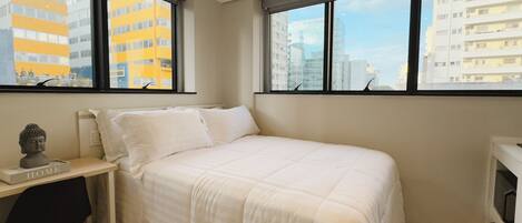Un dormitorio con una cama, un escritorio y una silla. Amplias ventanas con vista a la ciudad.