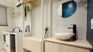 A modern bathroom with a white sink, black faucet, and a round mirror. There is a wooden cabinet below the sink and a small plant on the floor.